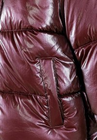 Giacca a vento bordeaux con finitura lucida, cuciture in vista e una tasca laterale. Design compatto per calore e copertura.