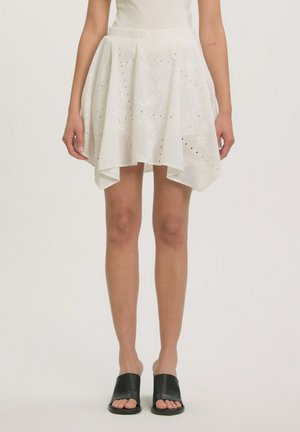 Jupe blanche à taille haute avec des détails en œillets et broderies florales, portée avec des chaussures noires ouvertes à enfiler sur une personne debout.