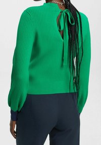 Grön ribbad tröja med långa puffärmar, rund halsringning och knytning i ryggen. Bärs med mörkblå byxor. Mjuk textur och figurnära design.