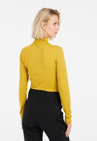 Dlouhý rukáv, přiléhavý top v hořčicově žluté barvě, s vysokým límcem a hladkým povrchem, spojený s černými kalhotami s vysokým pasem.