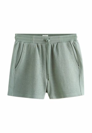 Person, die hellgrüne Shorts mit Kordelzug anpasst, getragen mit einem weißen gerippten Tanktop und einem grauen Pullover, der über die Schultern gelegt ist.
