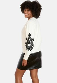 Maglione crema con ricami floreali neri su una manica, abbinato a una gonna di pelle lucida nera. Il maglione ha un collo rotondo e polsini a coste.
