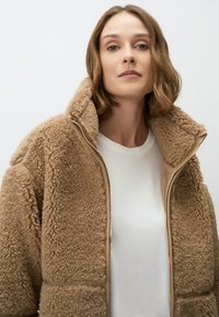 Bruine nepleren jas met hoge kraag en rits aan de voorkant, gecombineerd met een eenvoudige witte t-shirt. Zachte textuur en oversized ontwerp.