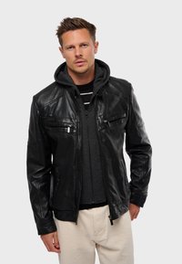 Chaqueta de cuero negro con cierre frontal y dos bolsillos en el pecho, superpuesta a una sudadera gris con capucha. Terminada con un corte ajustado y una textura suave.