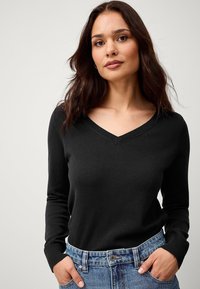 Maglione nero a V in maglia a coste, stile attillato, maniche lunghe, indossato sopra pantaloncini di jeans a vita alta con cuciture visibili.