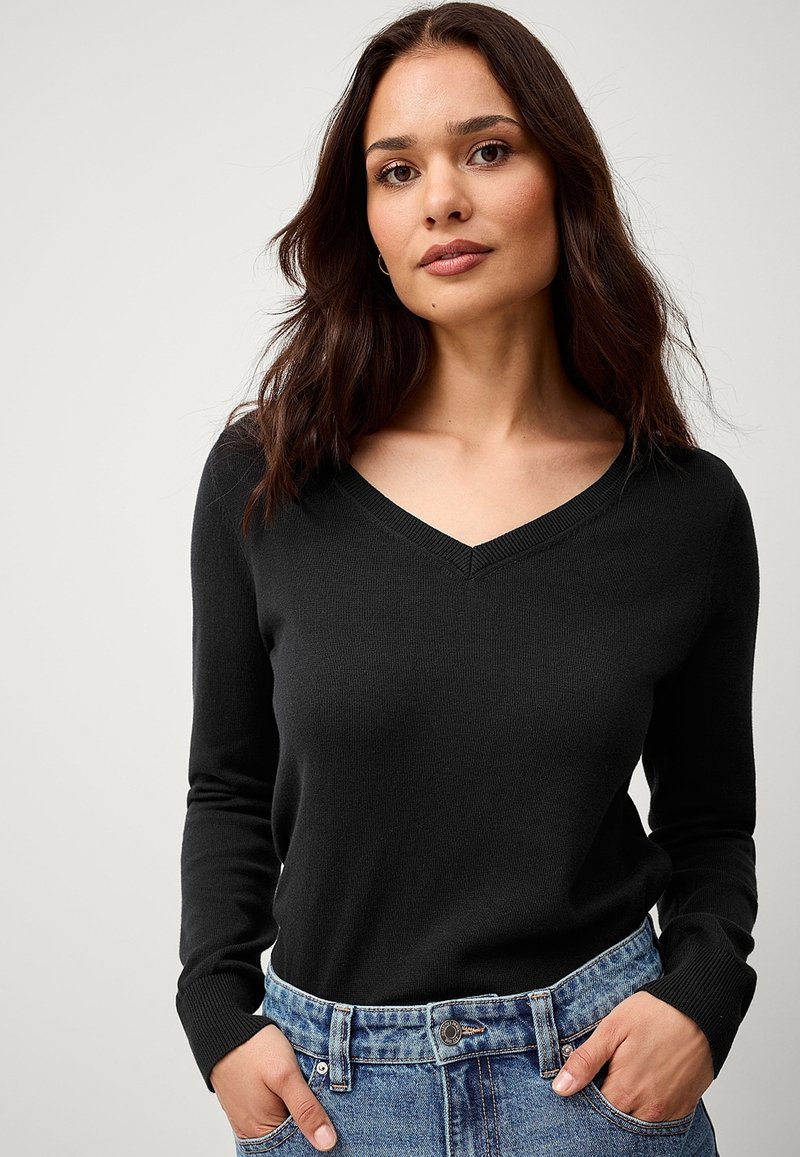 Maglione nero a V in maglia a coste, stile attillato, maniche lunghe, indossato sopra pantaloncini di jeans a vita alta con cuciture visibili.