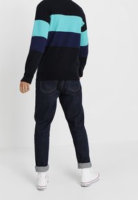 Strickpullover in Schwarz, Türkis und Marineblau mit horizontalen Streifen; kombiniert mit dunklen Jeans und weißen High-Top-Sneakern.