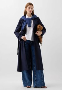Trench coat bleumarin, fabricat dintr-un material ușor, purtat peste o cămașă albă. Include blugi cu picioare largi și un pulover albastru aruncat pe umeri.