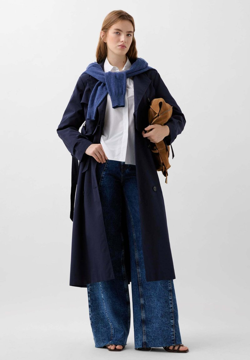 Trench coat bleumarin, fabricat dintr-un material ușor, purtat peste o cămașă albă. Include blugi cu picioare largi și un pulover albastru aruncat pe umeri.