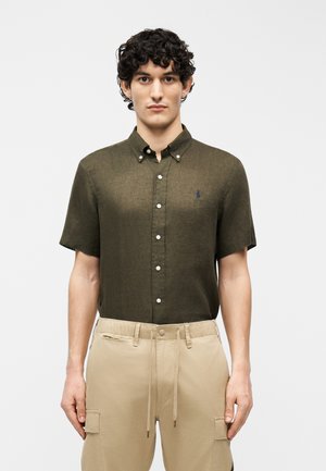 Man with curly hair wearing olive green short-sleeve button-up shirt and beige cargo pants, standing against plain background.