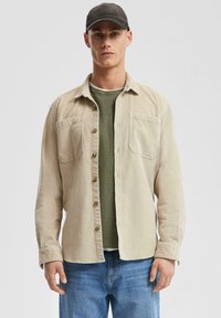 Beige corduroy overhemd met twee borstzakken, bruine knopen, gedragen over een donkergroen T-shirt, in combinatie met blauwe denim jeans.