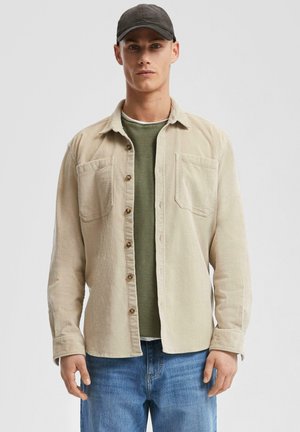 Beige corduroy shirt with two chest pockets, brown buttons, worn over a dark green T-shirt, paired with blue denim jeans.
