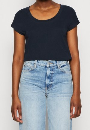 Osoba ubrana w prostą, ciemnogranatową bluzkę z krótkim rękawem i okrągłym dekoltem, włożoną w jasnoniebieskie dżinsy z wysokim stanem.