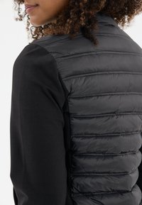 Svart pufferväst med horisontella sömmar, kombinerad med en svart långärmad topp. Den strukturerade materialet kontrasterar mot det släta tyget.