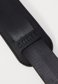 Cinghia nera in materiale sintetico con texture, caratterizzata dal logo "GUESS" impresso, superficie liscia e cuciture robuste lungo i bordi.