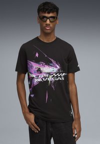 Joven con cabello rizado usando gafas de sol negras y una camiseta negra con un gráfico de un coche de carreras morado y blanco y el texto "LAS VEGAS".