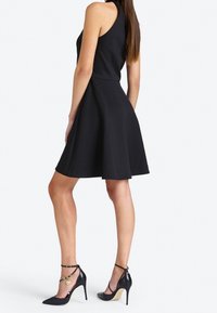 Robe noire sans manches avec un corsage ajusté et une jupe évasée, associée à des talons noirs à bout pointu avec une bride de cheville et des accessoires en or.