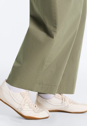 Weiße Schnürsneaker mit Gummisohlen, getragen mit weißen Socken und olivgrünen, weit geschnittenen Hosen, von den Knöcheln abwärts gezeigt.