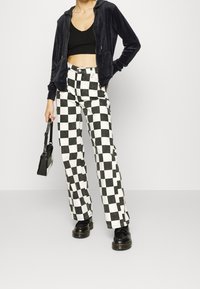 Personne portant un crop top noir, un sweat à capuche noir zippé, un pantalon à carreaux noir et blanc, des chaussures noires et tenant un petit sac à main noir.