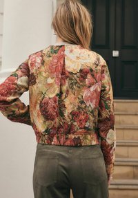 Kvinde med langt hår iført en cropped blomsterbroderet jakke og olivengrønne bukser, der står nær trapperne og en mørk dør.