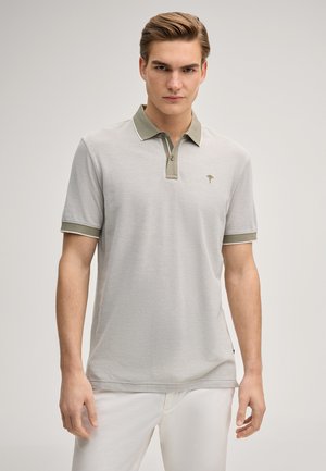 Młody mężczyzna w jasnoszarej koszulce polo z oliwkowozielonym kołnierzem i mankietami rękawów, stojący na tle jasnego, jednolitego tła.