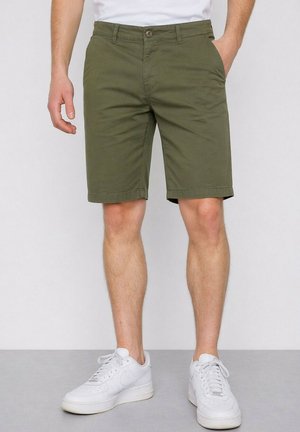 Homme portant un short vert olive au genou et des baskets blanches, debout devant un fond uni.