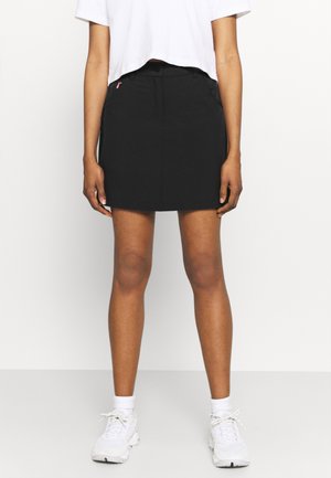 Persoon draagt een witte cropped top, zwarte minirok, witte sokken en witte vetersneakers en staat tegen een effen achtergrond.