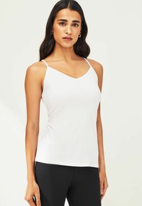 Witte mouwloze top met een V-hals ontwerp. Gemaakt van zachte stof, met dunne bandjes en een getailleerd silhouet. Zwarte legging wordt eronder gedragen.