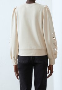 Persona che indossa un top crema a maniche lunghe con disegni floreali ritagliati sulle maniche e jeans neri, vista da dietro contro un muro semplice.