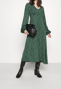 Grünes Kleid mit schwarzen Punkten, lange Ärmel, V-Ausschnitt, fließende Silhouette. Kombiniert mit einer schwarzen Lederhandtasche und knielangen schwarzen Stiefeln.