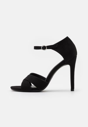 Black high-heeled sandals made of suede, featuring crossed straps on the toe and a thin ankle strap with a circular metal buckle.