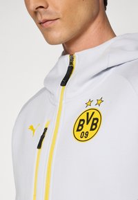 Valkoinen huppari, jossa on keltaisia yksityiskohtia, vetoketjullinen etuosa, Puma-logo ja Borussia Dortmundin joukkueen tunnus kahdella tähdellä. Pehmeä tekstuuri.