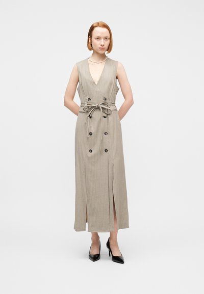 Γυναίκα στέκεται με τα χέρια πίσω από την πλάτη, φοράει αμάνικο μπεζ φόρεμα με κάθετες ρίγες, κουμπιά και ζώνη, μαύρα τακούνια και μαργαριταρένιο κολιέ.