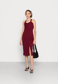 Vestido midi bordeaux com corpete ajustado, alças halter e tecido texturizado. Apresenta um corte lateral. Combinado com sandálias pretas de salto e uma mala.
