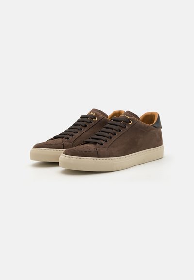 Sneaker in suede marrone con punta rotonda, suola in gomma di colore crema a contrasto e lacci scuri. Presenta un accento dorato sulla linguetta.