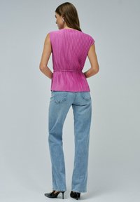 Mujer con una camiseta sin mangas de canalé rosa y cinturón, jeans rectos de mezclilla azul claro y tacones negros, de pie con la espalda hacia la cámara.