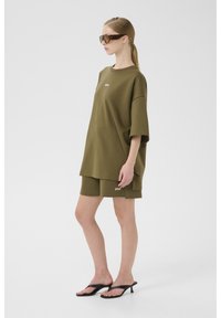 Olivnozeleni oversize majica in kratke hlače v mehkem materialu, z majhno logo, v kombinaciji s črnimi natikači in sončnimi očali s pisanimi vzorci.