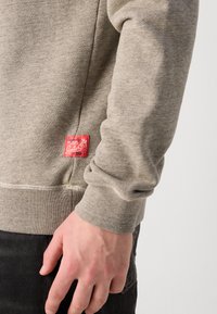 Lyse grå genser med ribbet tekstur, som har et rødt merke på siden og en avslappet passform. Ermet har en mansjett-design.