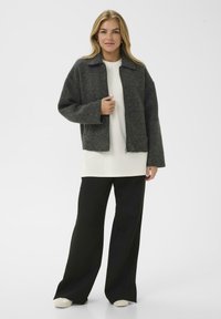 Junge Frau in einer dunkelgrauen Zip-Jacke, weißem Langarmoberteil, schwarzen weitem Hosen und weißen Schuhen, die vor einem schlichten Hintergrund steht.