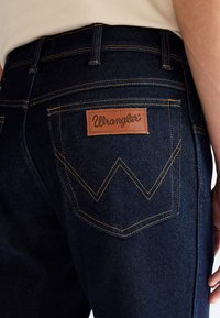Close-up of dark blue denim jeans back pocket with orange stitching and a brown leather Wrangler logo patch.