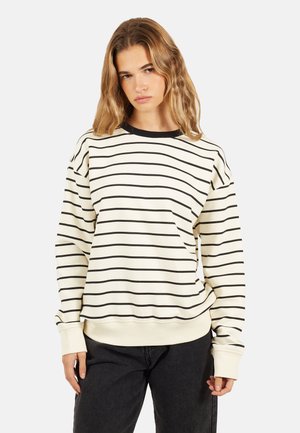 Giovane donna con capelli biondi mossi, che indossa un maglione bianco con strisce nere orizzontali e pantaloni neri, in piedi davanti a uno sfondo neutro.