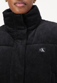 Sort fløjlskjorte med høj hals, quiltede sektioner og et cK-logo-patch på brystet. Den strukturerede stof øger det visuelle detaljer.