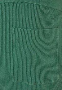 Zeleni rebrasti materijal s teksturiranim uzorkom koji sadrži veliku, zaobljenu džep na jednoj strani. Materijal izgleda mekano i izdržljivo.