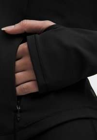 Jachetă neagră ajustată cu un buzunar cu fermoar; textură netedă, material elastic și detalii subtile de cusătură. Prim-plan al unei mâini în buzunar.