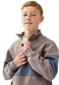 Ung pojke med fräknar som bär en grå och blå zip-up tröja, delvis dragande upp kragen, mot en vit bakgrund.