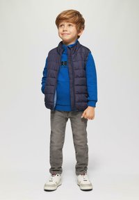 Blauwe sweatshirt met zwart logo, marineblauwe puffer vest, grijze jeans en witte sneakers. Jongen staat met ontspannen armen en glimlacht.