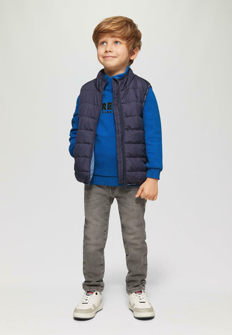 Blauwe sweatshirt met zwart logo, marineblauwe puffer vest, grijze jeans en witte sneakers. Jongen staat met ontspannen armen en glimlacht.