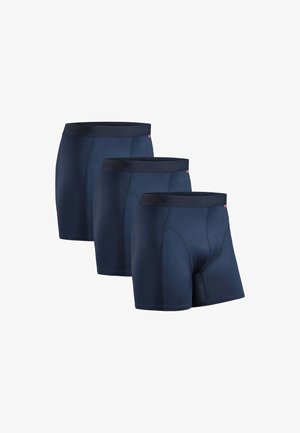 Tre marineblå boxershorts med sorte linninger arrangeret i et forskudt række på en hvid baggrund.