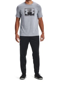 Šedé tričko s krátkými rukávy a velkým černým logem Under Armour. Kombinováno s černými sportovními kalhotami a černými botami, které mají texturovanou tkaninu.