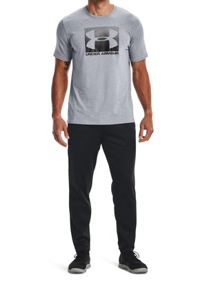 Szara koszulka z krótkim rękawem z dużym czarnym logo Under Armour. Dobraną parę stanowią czarne spodnie sportowe i czarne buty, wykonane z fakturowanej tkaniny.